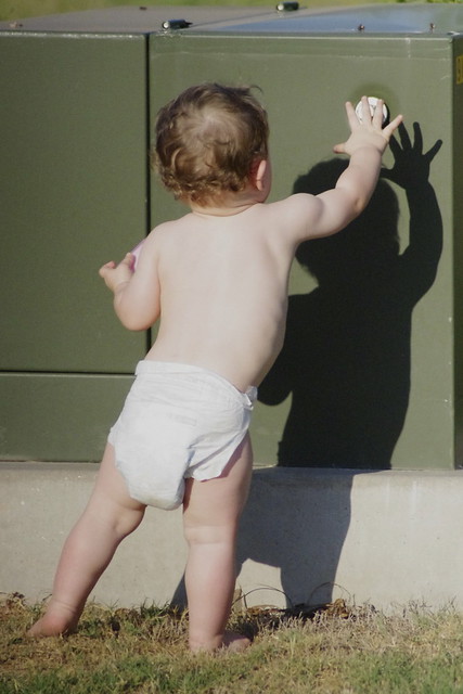 Fotografía de un bebé de espaldas, parado frente a una pared, jugando con su sombra.