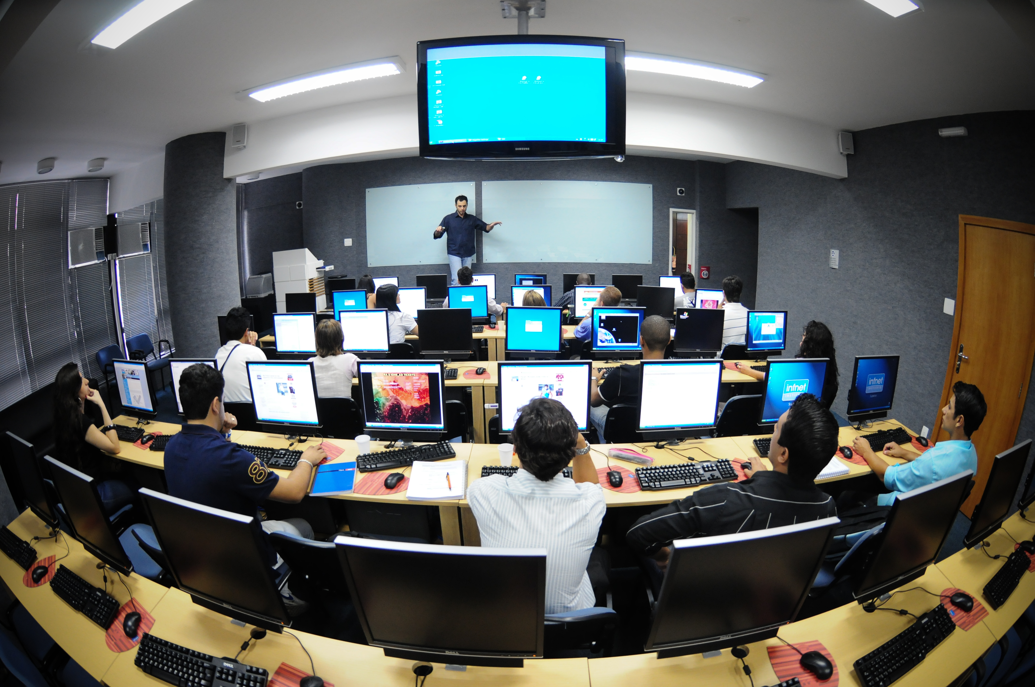 Aula con estudiantes usando computadoras y profesor al frente con pantalla digital 