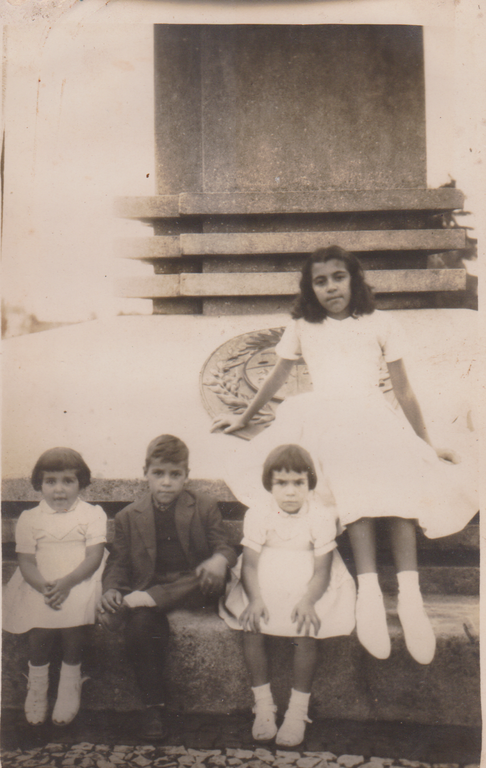A imagem contém o registro de crianças sentadas no Obelisco da Praça Internacional